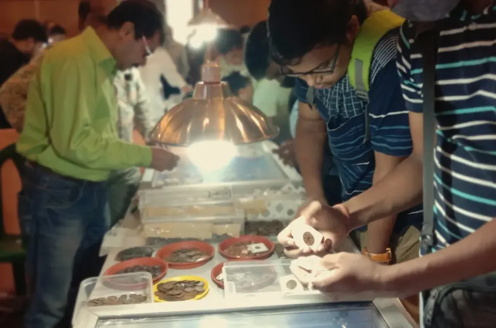 Men at a lit coin dealer table examining coins in holders above trays of loose coins