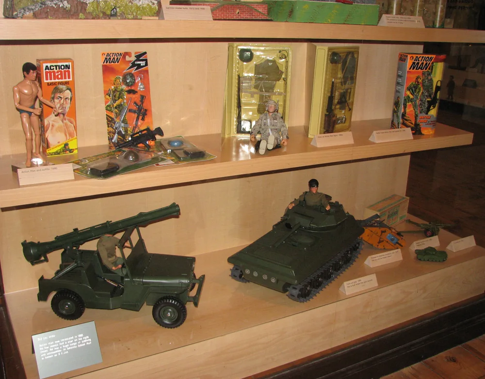 Museum display of vintage Action Man figures in original boxes, blister-carded accessory packs, a military jeep and a tank