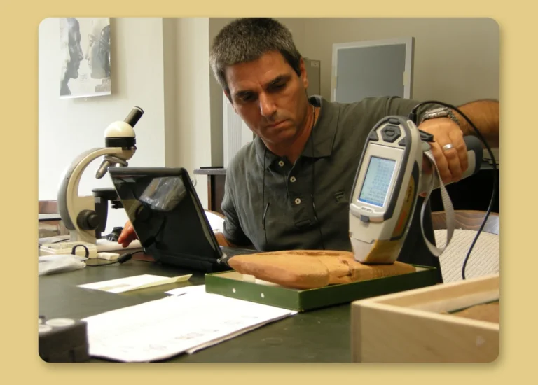 Researcher testing an object with a handheld X-ray fluorescence (XRF) analyzer, with a microscope on the desk